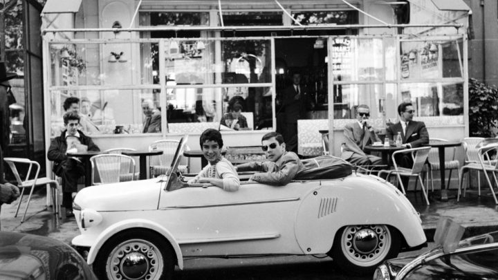 Deux personnes souriantes dans une petite voiture blanche ancienne devant un bar