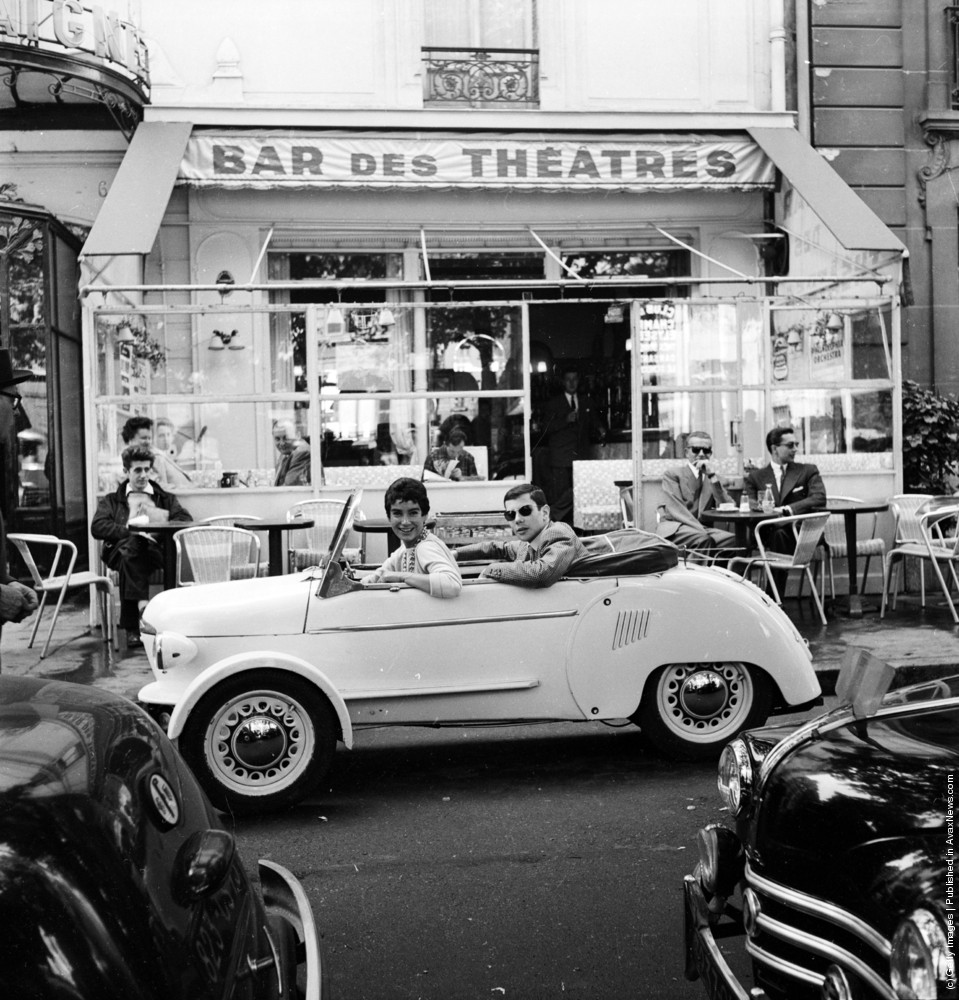 Deux personnes souriantes dans une petite voiture blanche ancienne devant un bar